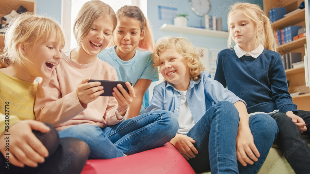 Diverse Group of Cute Small Children Sitting together on the Bean Bags Use Smartphone and Talk, Have Fun. Kids Browsing on Internet and Playing Online Video Games on Mobile Phone