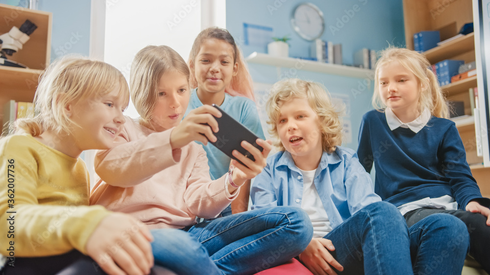 Diverse Group of Cute Small Children Sitting together on the Bean Bags Use Smartphone and Talk, Have Fun. Kids Browsing on Internet and Playing Online Video Games on Mobile Phone