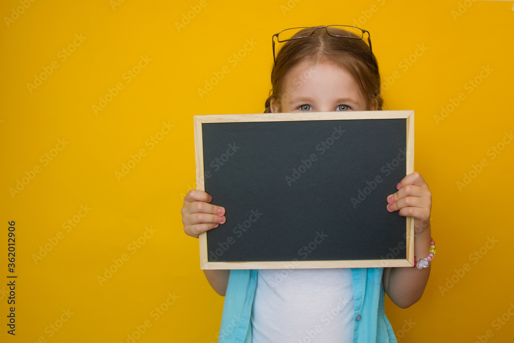 Portrait of a beautiful little girl peeking from behind a blackboard in her hands, smiling on a yellow background. Attractive positive baby. back to school. Copyspace Portrait of a beautiful little girl peeking from behind a blackboard in her hands, smiling on a yellow background. Attractive positive baby. back to school. Copyspace