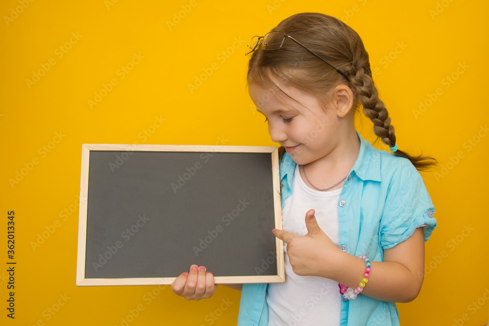 Back to school. A cheerful little girl with pigtails holds a school board in her hands with a place for text and points a finger at it on a yellow background. Copyspace Back to school. A cheerful little girl with pigtails holds a school board in her hands with a place for text and points a finger at it on a yellow background. Copyspace
