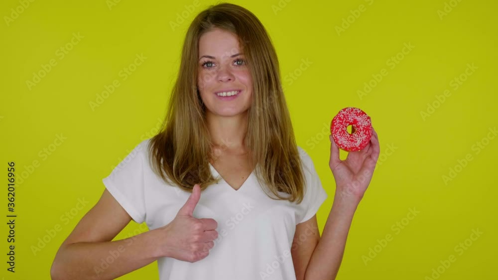 Cheerful woman in a white T-shirt raises hand with a red donuts, shows like and smiles on a yellow background with copy space. Place for text or product. 4K slow motion footage Cheerful woman in a white T-shirt raises hand with a red donuts, shows like and smiles on a yellow background with copy space. Place for text or product. 4K slow motion footage