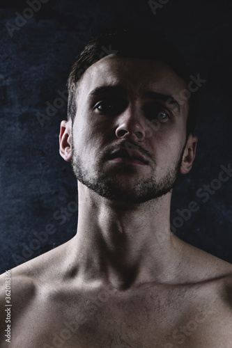 Art portrait of an emotional young unshaven brunette man with a serious face and a bare torso against a dark wall