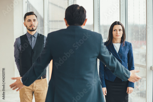 Fotografie Angry boss standing back to the camera, shouting at his workers, raising hand, while stressed employees standing, with guilty look on their faces