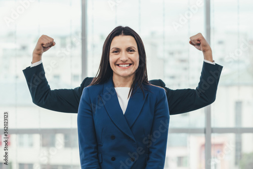 Beautiful businesswoman stands, looking at the camera smiling, while funky man plays around, standing behind her, so that only his hands raised up are seen.