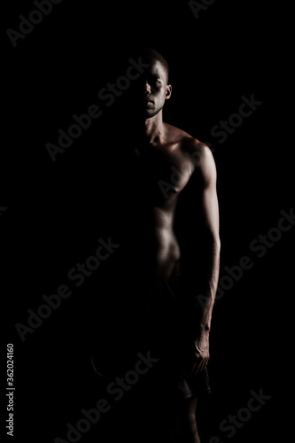 Bare chested black fitness man silhouette looking at camera wearing sport short trousers on black backgorund