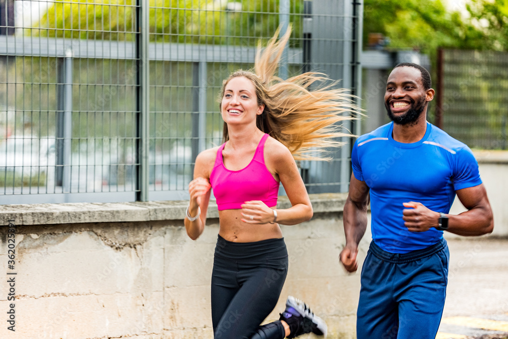 Young couple smiling to each other while jogging on the street, out to run. Afro american man and beautiful blonde girl in sportswear running through the city street together 