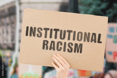 The phrase " Institutional racism " on a banner in men's hand with blurred background. Inequality. Social issues. Discrimination. Human rights. Community. Relationship. Politics. Injustice