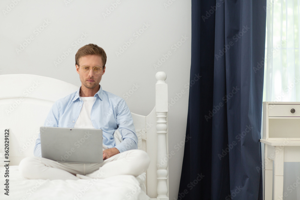 Guy working with laptop in his bed at home. Calm peacefull middle aged man wearing pyjamas and thin clear glasses sitting in his bed under blanket and working with computer.