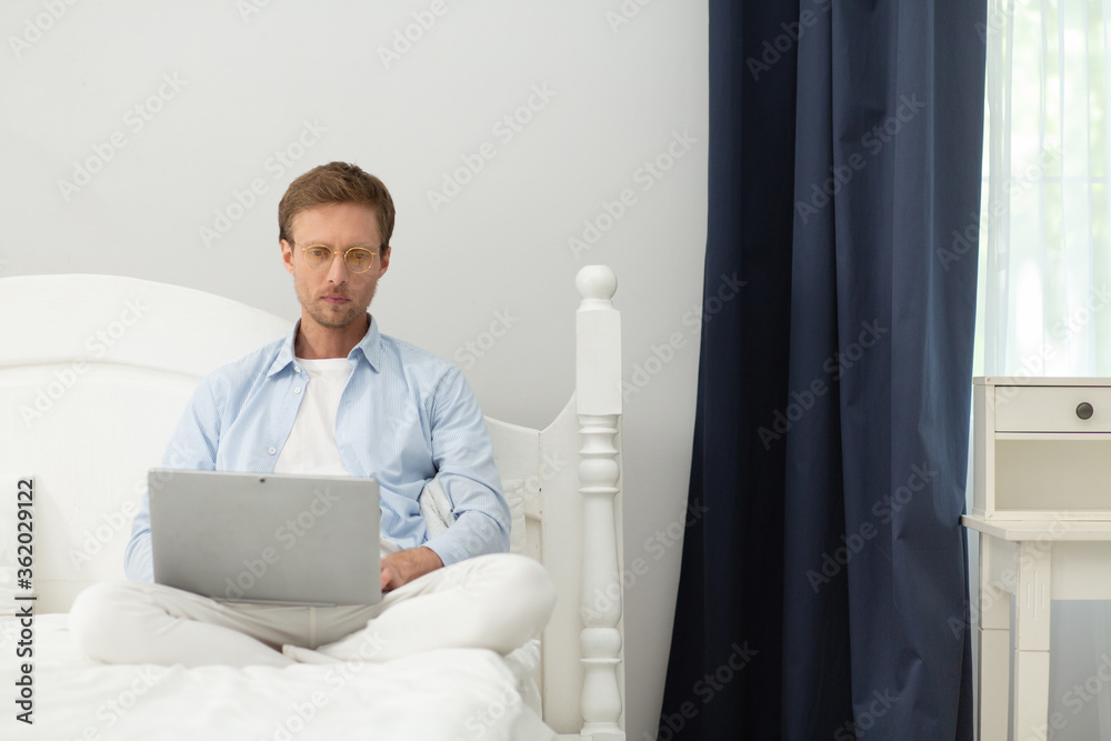 Guy working with laptop in his bed at home. Calm peacefull middle aged man wearing pyjamas and thin clear glasses sitting in his bed under blanket and working with computer. Guy working with laptop in his bed at home. Calm peacefull middle aged man wearing pyjamas and thin clear glasses sitting in his bed under blanket and working with computer.