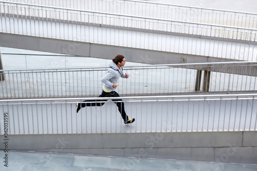 Full length portrait of handsome runner jogging fast down the bridge in urban setting with copy space area for your text message or content, young male jogger working out outdoors on a cloudy day