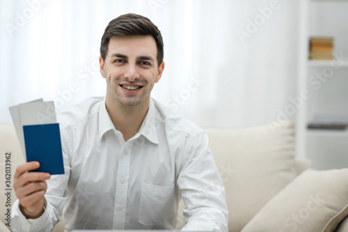 Closeup of man holding tickets for plane and passport ready for travelling, sitting on the sofa at home.