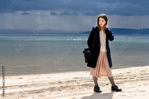A young adult beautiful girl stands on the shore against the backdrop of an approaching cloud with rain. The woman is wearing a spotty dress, a white sweater and a navy coat.