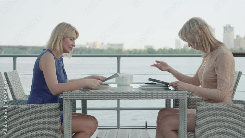 Side view wide shot of confident businesswomen discussing menu in outdoor restaurant. Portrait of successful Caucasian women resting in luxurious cafe on river bank. Luxury leisure, lifestyle. Side view wide shot of confident businesswomen discussing menu in outdoor restaurant. Portrait of successful Caucasian women resting in luxurious cafe on river bank. Luxury leisure, lifestyle.
