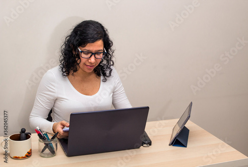 Young black-haired Caucasian woman in a white shirt and glasses works from home with her laptop and tablet