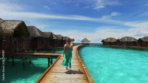 Maldive island resort. Girl in green dress walking on wooden bridge near bungalow hotel. View of a young woman with long hair from the back. Maldives travel destination. Summer vacation in paradise