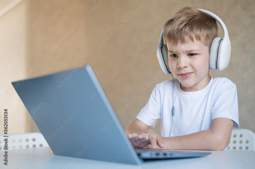 Cute little boy is watching cartoons or a movie on a laptop. Caucasian child sits at a table at home and listens to music on headphones. Cute little boy is watching cartoons or a movie on a laptop. Caucasian child sits at a table at home and listens to music on headphones.