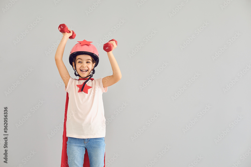A child in a costume of a super hero raised his hands up with dumbbells on a gray background. A child in a costume of a super hero raised his hands up with dumbbells on a gray background.