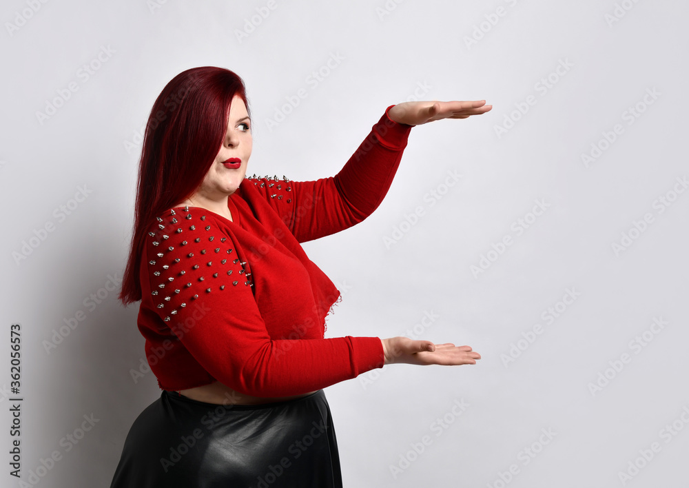 Plus size ginger girl in red spiked top, black leather skirt. Looking surprised, holding something big, posing isolated on white Plus size ginger girl in red spiked top, black leather skirt. Looking surprised, holding something big, posing isolated on white