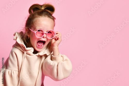 Naughty little girl with tall hair dressed in pink hoodie yelling angrily resettling spectacles on face. Close up shot isolated on pink  copy space