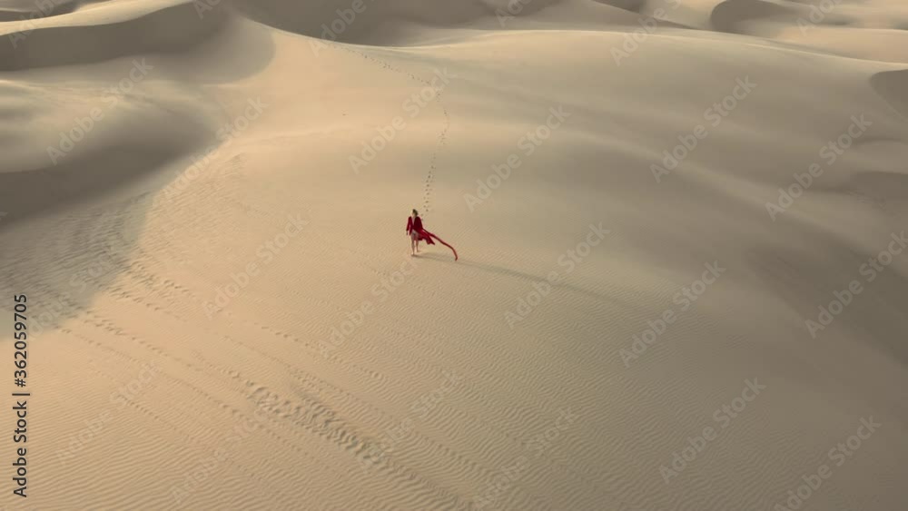 A lonely traveler in fashion red fluttering dress is walking by the beige sand dunes. 4K aerial remote view of a beautiful female model in the desert at sunset, California. Nature landscape footage
