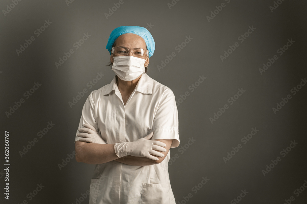 Doctor crossed her arms looking at camera. Female caucasian medic in protective mask and white uniform stands on gray background. Empty space for text at right. Tinted image. Doctor crossed her arms looking at camera. Female caucasian medic in protective mask and white uniform stands on gray background. Empty space for text at right. Tinted image.