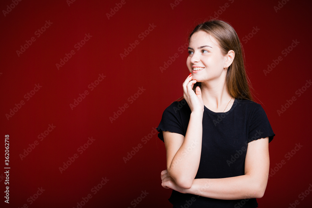 Fototapeta premium Beautiful American woman in a black T-shirt holds a hand near her face and looks away smiling on a red background