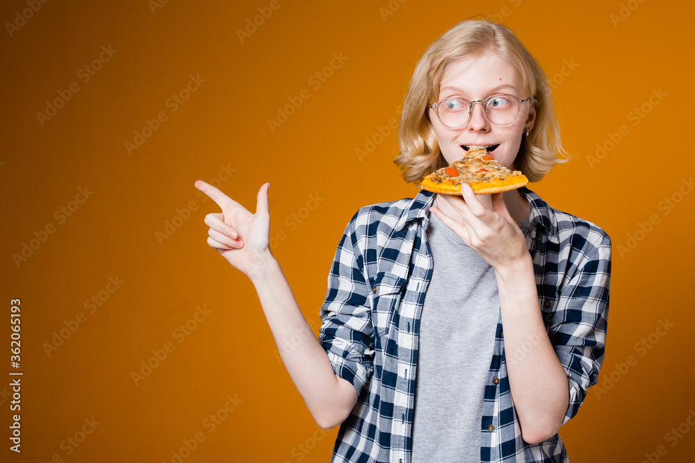 A girl of European appearance holds a piece of pizza at her mouth and points her finger to the side to an empty place to insert her text on an orange background A girl of European appearance holds a piece of pizza at her mouth and points her finger to the side to an empty place to insert her text on an orange background