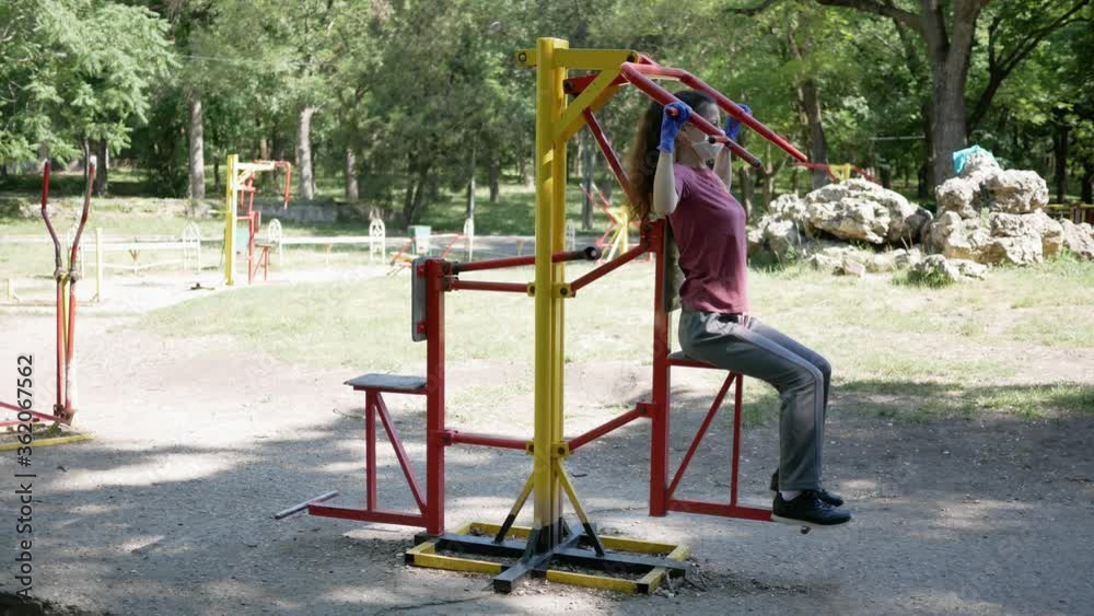 Young woman in gloves and face mask sports on outdoor exercise equipment in Park. Protection from epidemic of coronavirus in use of simulators in public places Young woman in gloves and face mask sports on outdoor exercise equipment in Park. Protection from epidemic of coronavirus in use of simulators in public places