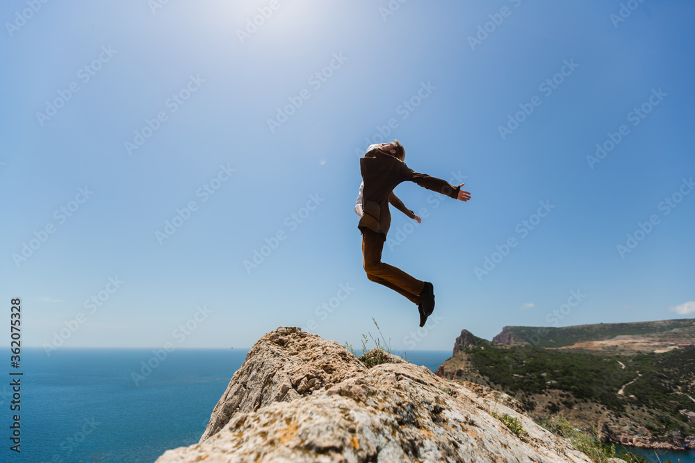 The silhouette of a young man in a brown suit, standing on the edge of a cliff and jumping in the pose of a flying bird with his hands divorced against the bright blue sky and scorching sun