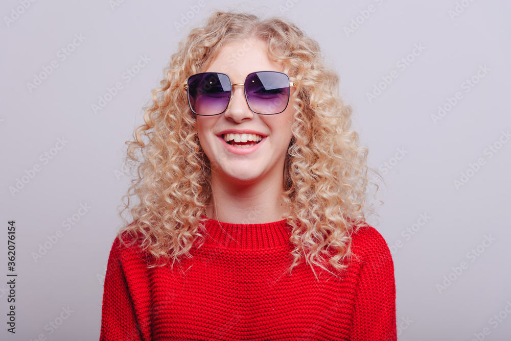 Portrait of a cheerful smiling young girl in glasses with curly blond hair in a studio wearing a red sweater.