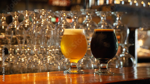 Two full beer glasses, on a counter at a bar, a stack of glasses and beer taps in the background, reopening after covid