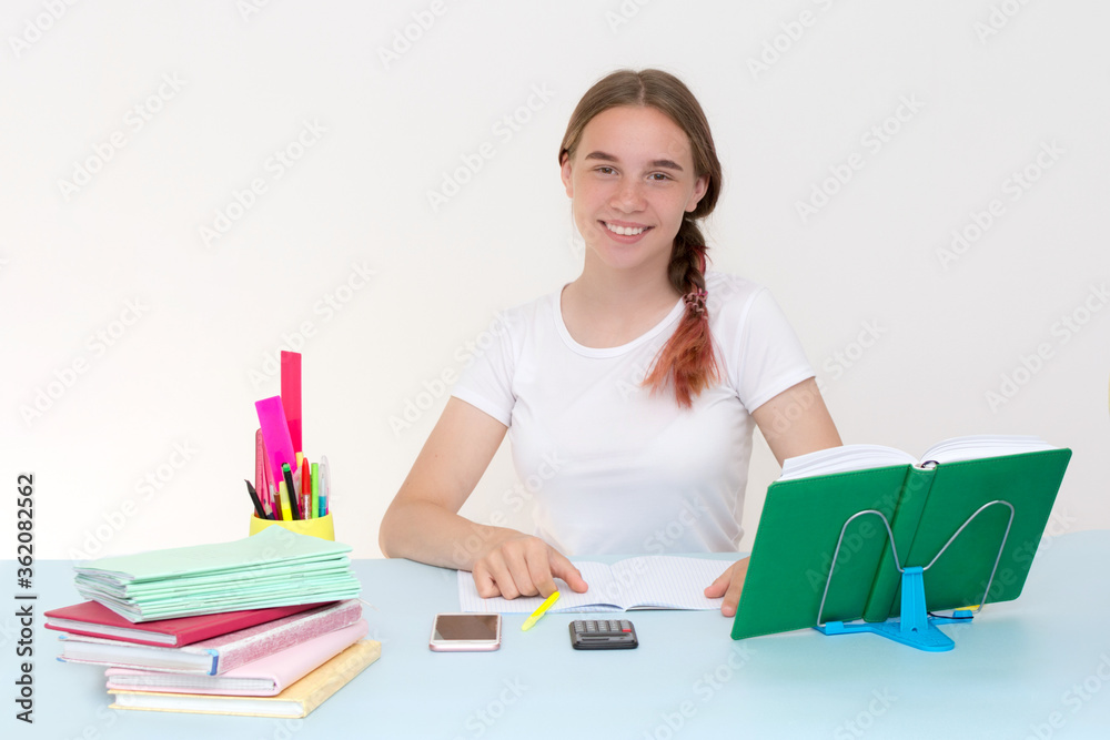 A beautiful teenager girl sits at a table on which lie notebooks, textbooks, a calculator, a phone. Schoolgirl. She is looking into the frame, smiling. Horizontal photo. A beautiful teenager girl sits at a table on which lie notebooks, textbooks, a calculator, a phone. Schoolgirl. She is looking into the frame, smiling. Horizontal photo.