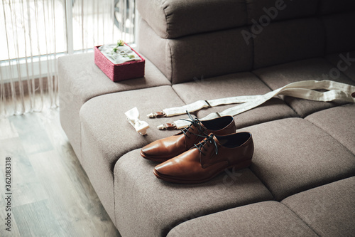 Men's accessories outfits. Brown formal leather shoes, bow-tie and suspenders prepared for dressing. Elements of groom's clothes laying on sofa.
