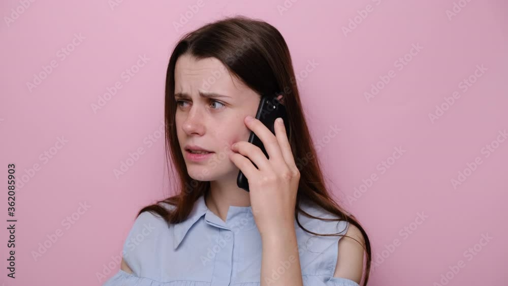 Close up portrait of angry young caucasian woman talking on smartphone, dressed in blue shirt, isolated over pink wall background in studio. People, lifestyle, emotions and stress concept Close up portrait of angry young caucasian woman talking on smartphone, dressed in blue shirt, isolated over pink wall background in studio. People, lifestyle, emotions and stress concept