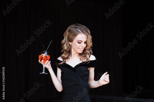 Beautiful blonde, young girl holding an aperol spritz. Aperol cocktail syringe in a glass. Portrait of a young girl in a restaurant. Young girl in a restaurant with a glass on a black background.