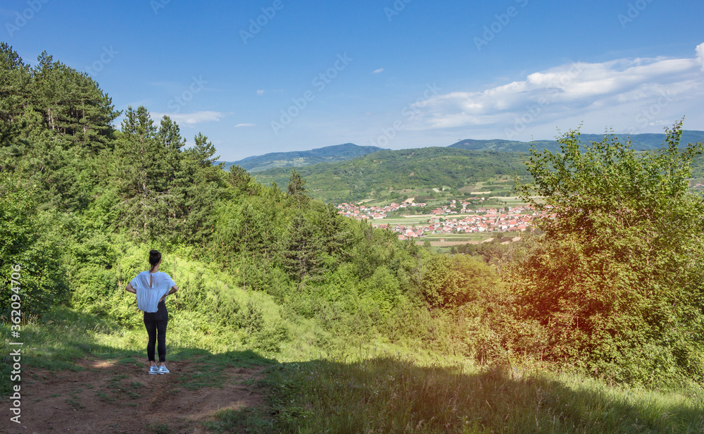 Back view of young woman standing on top of a hill. Attractive girl enjoying the colorful countryside view. Small village at the foot of a hill, beautiful landscape with blue sky.