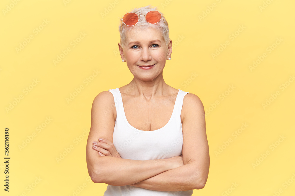 Beautiful Caucasian retired woman wearing round pink sunglasses and white tank top, crossing arms on her chest, having confident facial expression. People, age, confidence and lifestyle concept Beautiful Caucasian retired woman wearing round pink sunglasses and white tank top, crossing arms on her chest, having confident facial expression. People, age, confidence and lifestyle concept