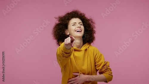 Hey you! Amused girl afro hairstyle in hoodie pointing finger to camera, laughing out loud, taunting making fun of ridiculous appearance, funny joke. indoor studio shot isolated on pink background photo
