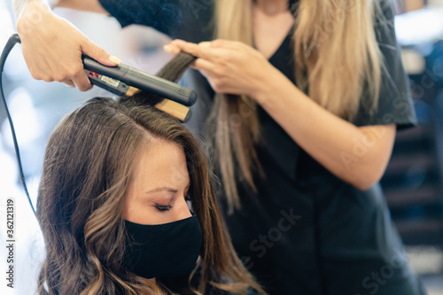 Hairdresser, protected by a mask, combing her client's hair with a hair iron in a salon.
