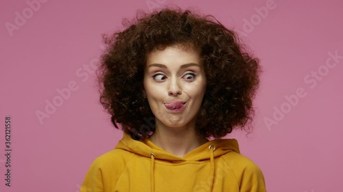 Funny amusing girl afro hairstyle in hoodie looking awkward with crossed eyes and puffed cheeks, fooling around making stupid brainless dumb expression. indoor studio shot isolated on pink background