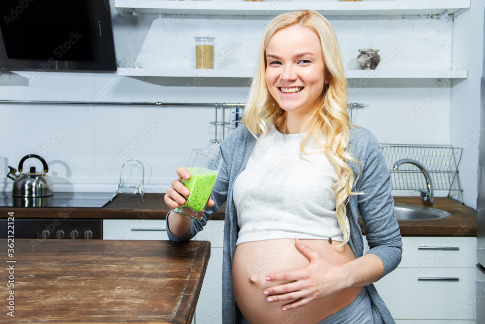 Young pregnant woman is drinking healthy organic green smoothie on kitchen at home. Girl is touching, stroking her tummy. Nutrition and care during pregnancy. Expectation of baby concept. Young pregnant woman is drinking healthy organic green smoothie on kitchen at home. Girl is touching, stroking her tummy. Nutrition and care during pregnancy. Expectation of baby concept.