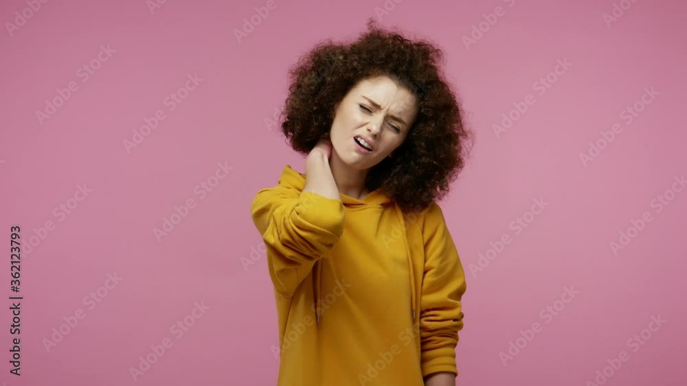 Unhealthy tired girl afro hairstyle in hoodie touching sore neck feeling fatigued, doing massage, warming up painful stiff muscles, hurting shoulders. indoor studio shot isolated on pink background Unhealthy tired girl afro hairstyle in hoodie touching sore neck feeling fatigued, doing massage, warming up painful stiff muscles, hurting shoulders. indoor studio shot isolated on pink background