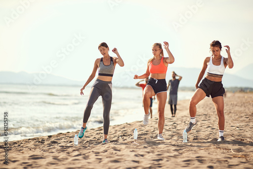 group of three women exercise on a beach with two men walking in the background