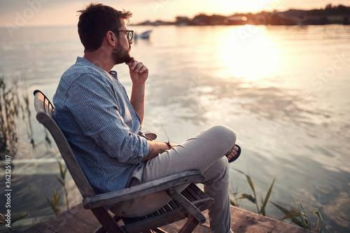 Fotografie, Obraz beardy guy sitting alone on a river coast, enjoying the sunset, thinking