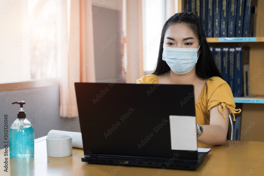 A teenager student wears surgical mask and studies online via laptop during COVID-19 pandemic. University student girl watches online classes and writing a syllabus in notebook. Stock photo. A teenager student wears surgical mask and studies online via laptop during COVID-19 pandemic. University student girl watches online classes and writing a syllabus in notebook. Stock photo.