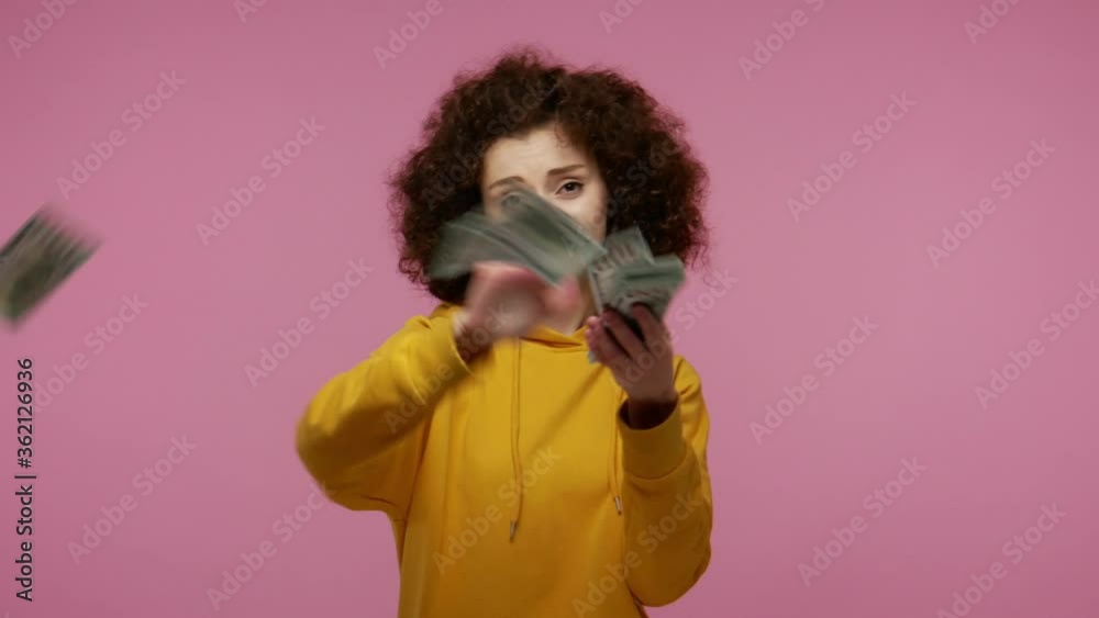Wasteful girl afro hairstyle in hoodie scattering dollars looking arrogant, boasting wealthy life, concept of careless money spending, squandering throwing around cash. indoor studio shot isolated Wasteful girl afro hairstyle in hoodie scattering dollars looking arrogant, boasting wealthy life, concept of careless money spending, squandering throwing around cash. indoor studio shot isolated