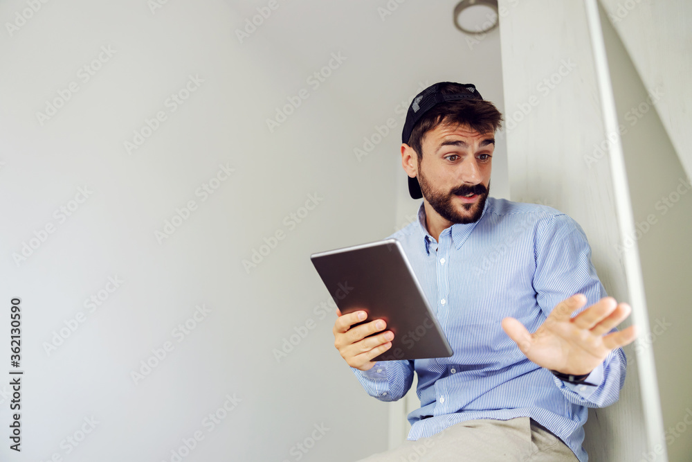 Young handsome businessman dressed business casual sitting in office and using tablet while talking to colleague.