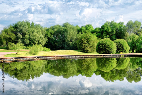 picturesque countryside landscape on morning against blue clouds sky background. Wide view of pond embankment at park in Moscow Russia with mirror reflection of scenery in water. Summer rural scene