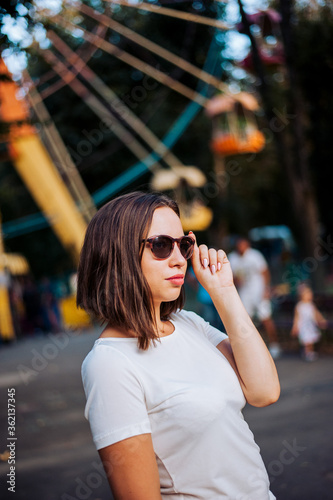 girl with glasses in the о background of the Ferris wheel in the amusement park. The girl looks at the camera and takes off her glasses. Girl in white T-shirt walks in the park
