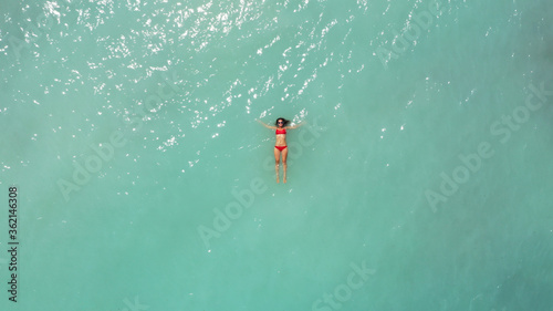 Aerial view of young beautiful woman in red bikini and sunglasses lying on light blue azure turquoise water of sea, relaxing in ocean. Summer beach vacation. Malibu lifeguard. Maldives. Melasti beach.